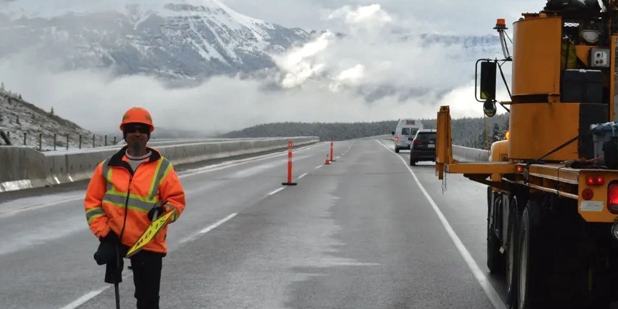 Road worker in high-visibility gear on a mountain highway with snow-covered peaks.