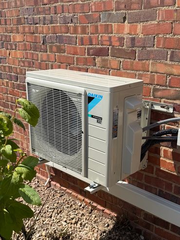 Outdoor Daikin air conditioning unit mounted on a brick wall with greenery nearby.