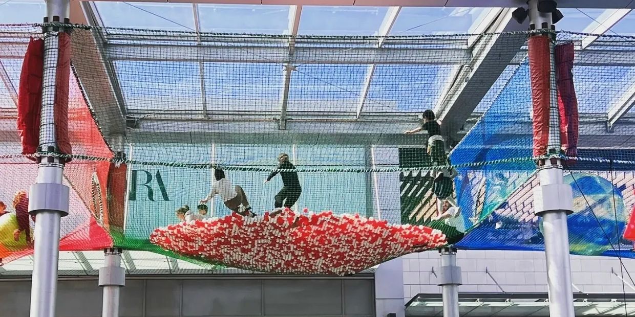Children playing on a suspended net filled with red and white balls under a glass roof.
