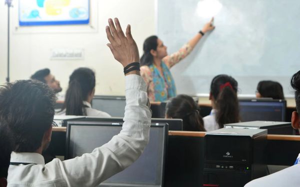 Student raising hand in computer classroom while teacher points at whiteboard.