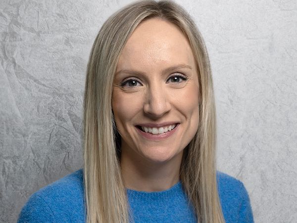 Smiling woman with straight blonde hair wearing a blue sweater against a textured gray background.