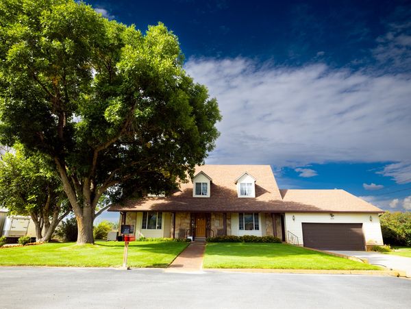 A suburban house with a large tree and a well-maintained lawn under a partly cloudy sky.