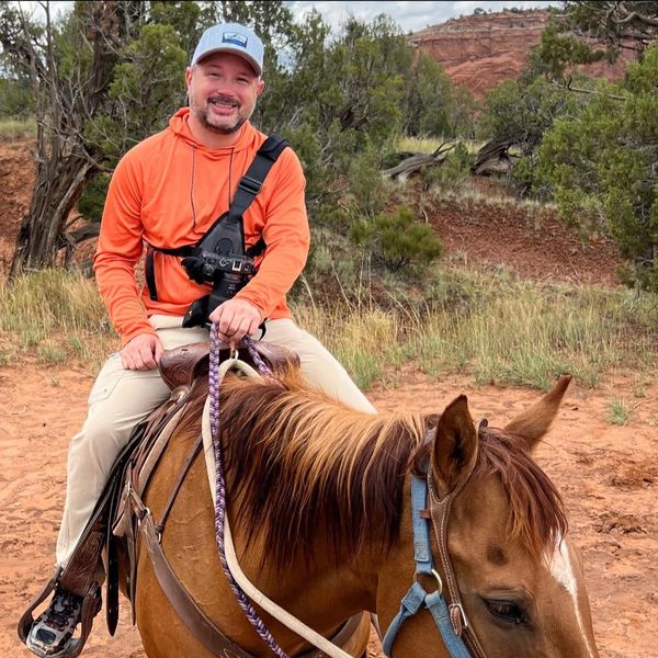 A man in an orange hoodie rides a brown horse in a natural, rocky landscape.