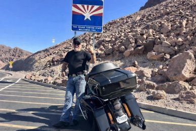 Man poses with motorcycle near Arizona state welcome sign in rocky desert.