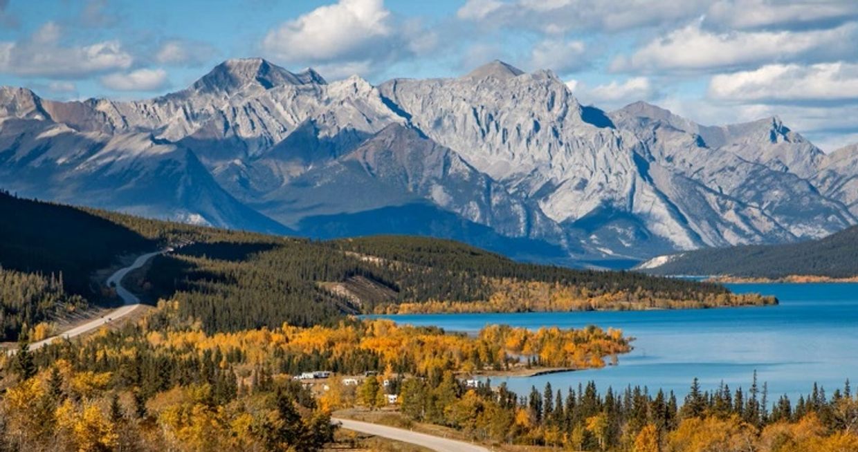 Scenic mountain range beside a lake with autumn trees and a winding road.