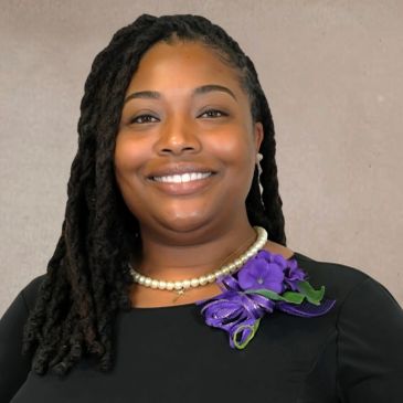 Smiling woman with braided hair, wearing a pearl necklace and purple floral corsage.