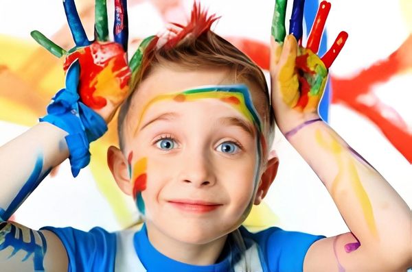 A joyful child with blue eyes and colorful paint on face, hands, and hair looks at the camera.