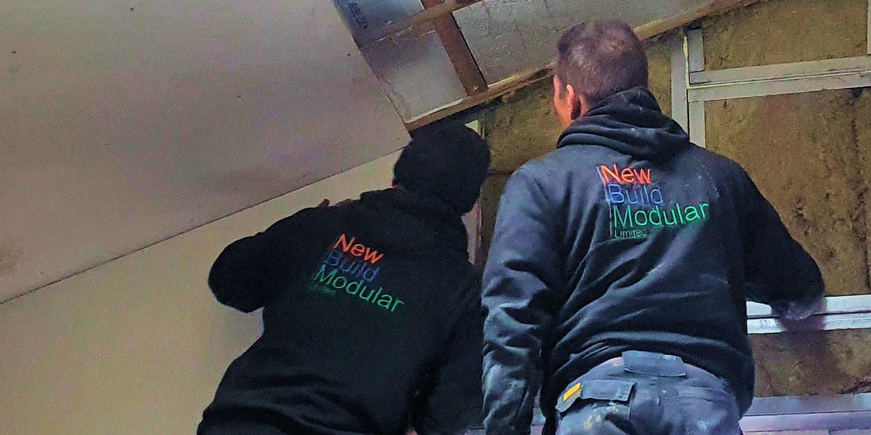 Two workers installing ceiling panels wearing New Build Modular hoodies.