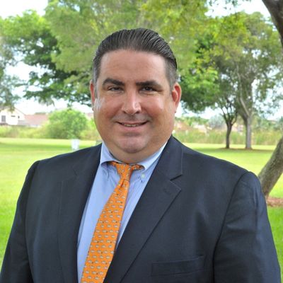 Man in a suit with an orange tie standing outdoors in a park.