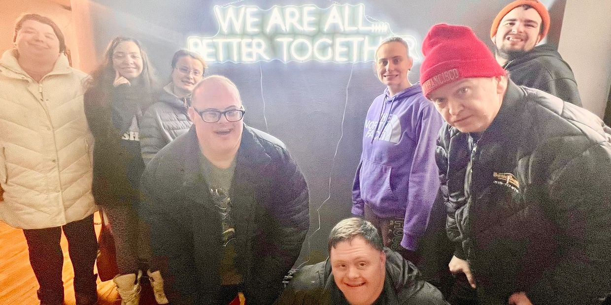 A diverse group of people smiling under a neon sign that reads 'We are all better together.'