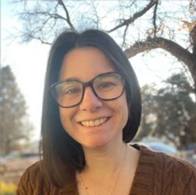 Smiling woman with glasses and brown sweater outdoors in soft sunlight.