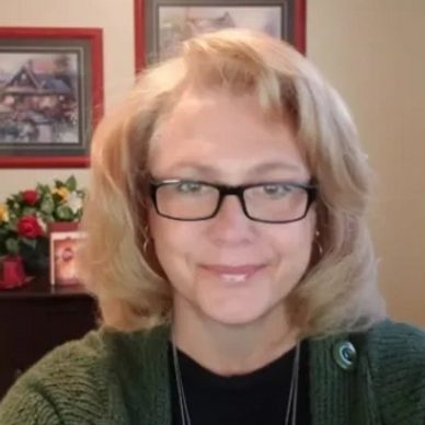Smiling woman with glasses and blonde hair indoors, wearing a green sweater.