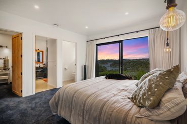 Cozy bedroom with a large window showing a colorful sunset and a dog resting on the bed.