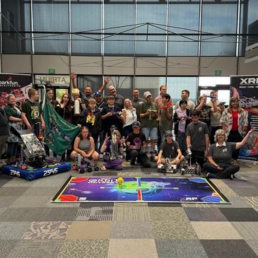 Group photo of a robotics team with robots and banners in a large indoor space.