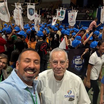 A lively group of people wearing blue helmets at a robotics event, with three adults smiling in the foreground.