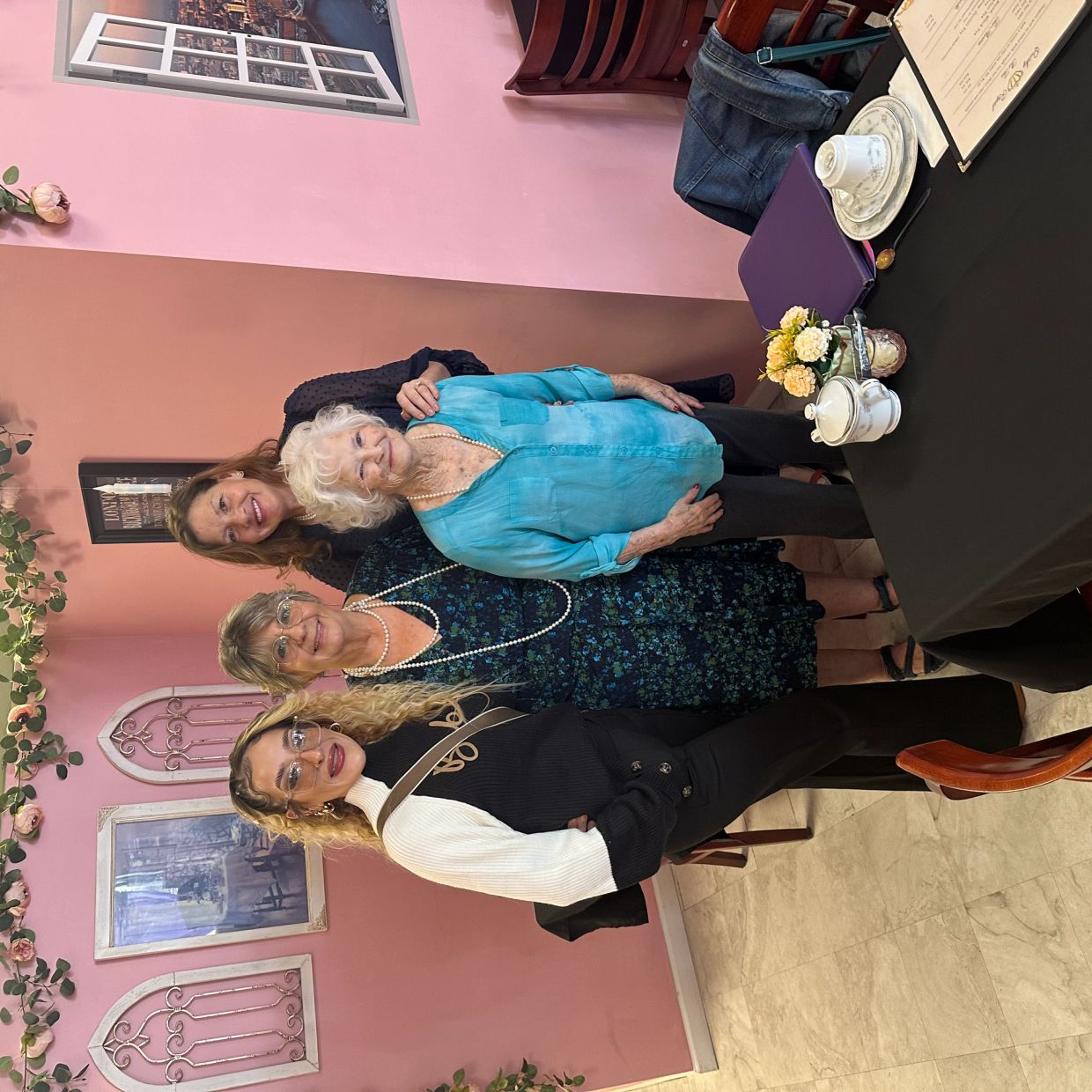 Four women posing together in a cozy, flower-decorated room with pink walls.