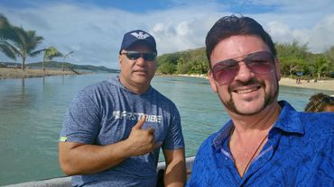 Two men smiling by the water with palm trees in the background.