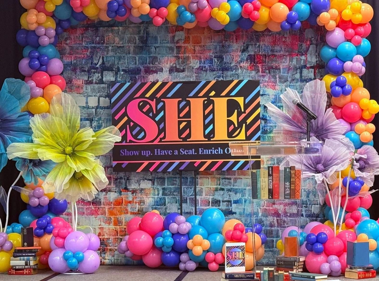 Colorful SHE event stage with balloons, large flowers, and books.