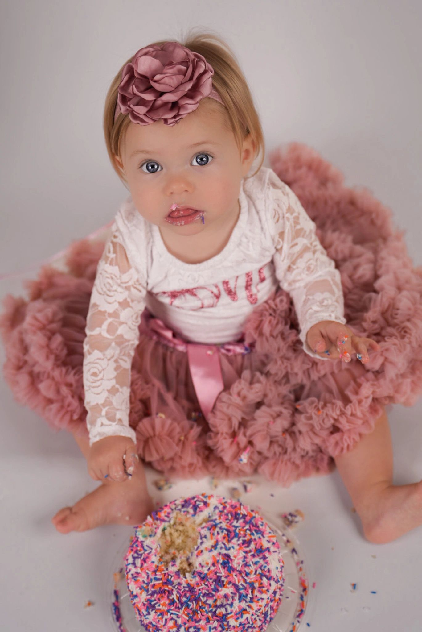 Baby girl in a pink dress enjoying a cake with colorful sprinkles.