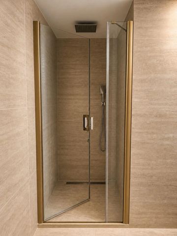 Modern shower with glass door and gold trim in beige tiled bathroom.