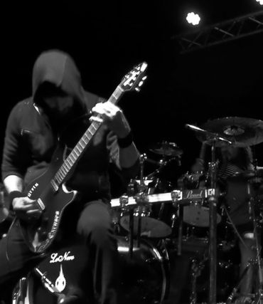 Musician in a hoodie playing electric guitar on stage with a drummer in the background.
