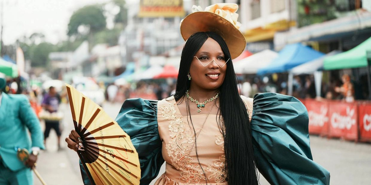 Woman in elegant vintage dress with a fan and hat smiles at a street event.