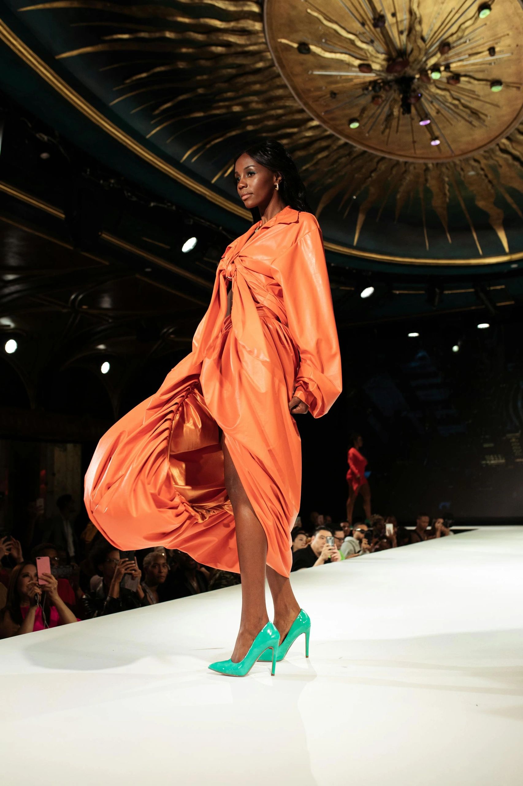 Model in a flowing orange dress and teal heels on runway.