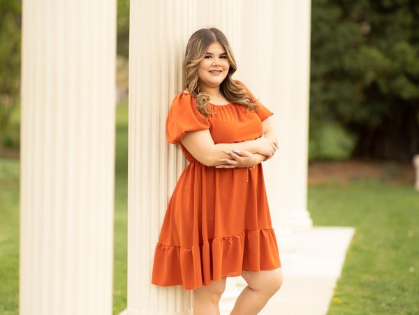 A woman in an orange dress leaning against a white column outdoors.
