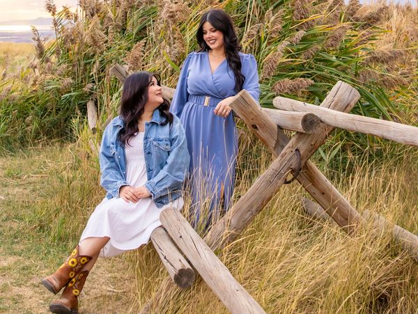 Two women smiling outdoors by a rustic wooden fence surrounded by tall grass.