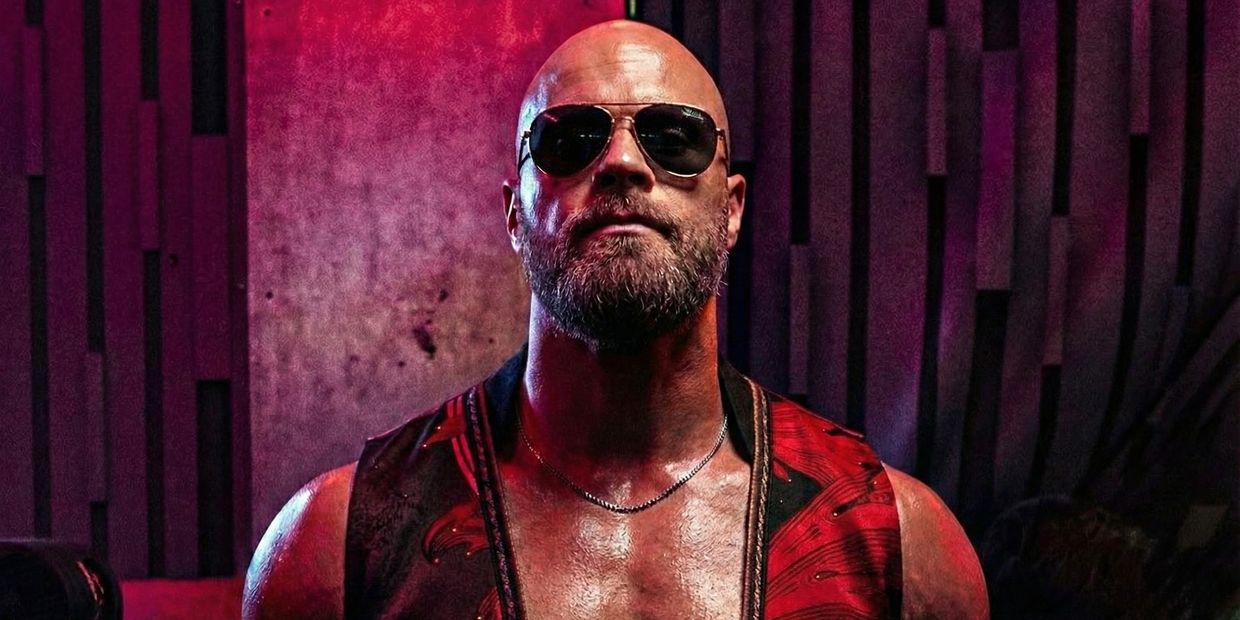 Bald man with beard wearing sunglasses and a red patterned vest in dramatic lighting.
