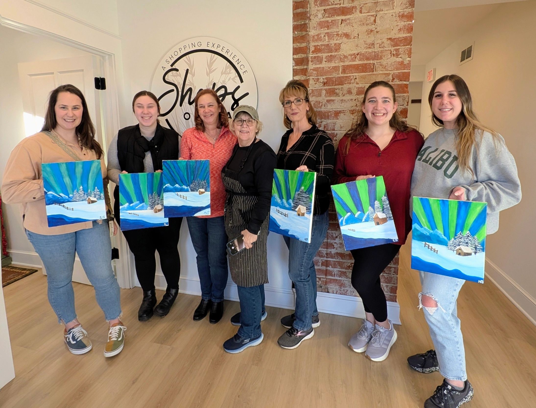 Group of women holding winter landscape paintings, smiling indoors.