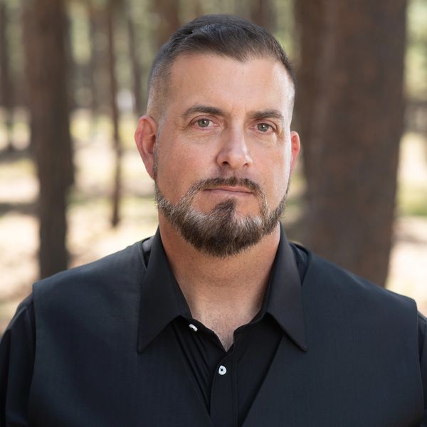 Man with a beard wearing a black shirt and vest outdoors in a forest.