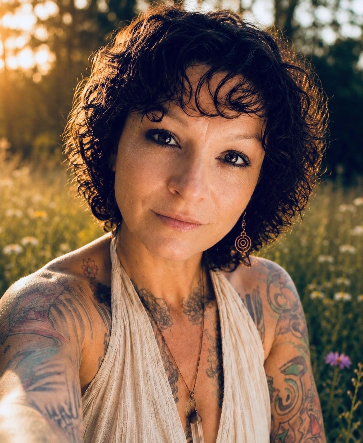 Woman with curly hair and tattoos posing outdoors at sunset.
