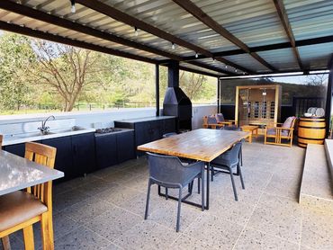 Covered outdoor braai area with built-in grill, sink, wooden dining table, seating, & lounge chairs