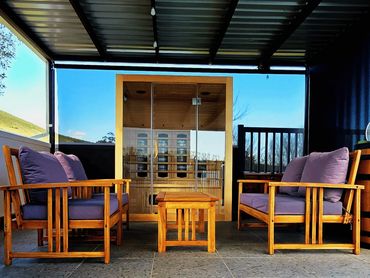 Cozy outdoor lounge with wooden chairs and purple cushions facing a modern glass-front sauna