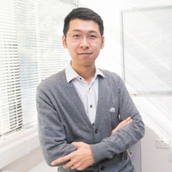 Man in glasses and gray cardigan standing with arms crossed indoors.