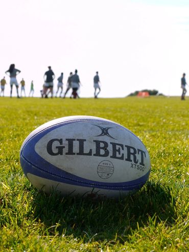 A Gilbert rugby ball sits in the grassy foreground as women's players practice in background