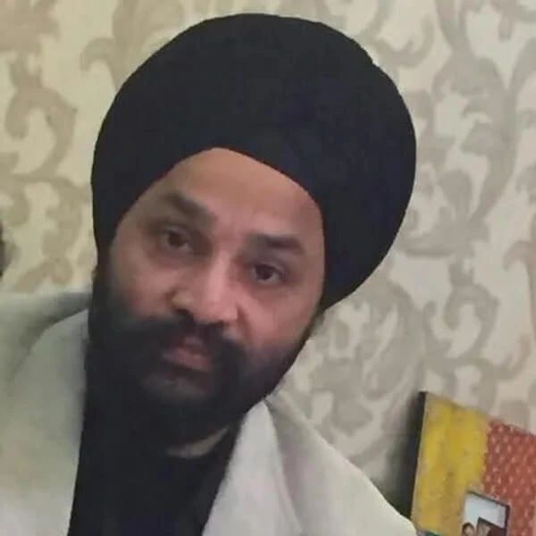 Man wearing a black turban and light-colored coat in front of a patterned wall.