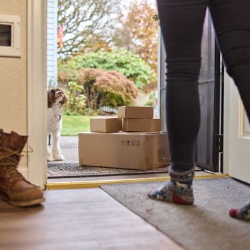 Woman opening her front door to delivery boxes with a dog peeking in, autumn leaves outside.