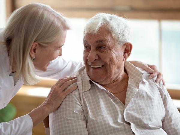 Middle aged care female licensed practical nurse in white coat talk to patient aged 80 years old man