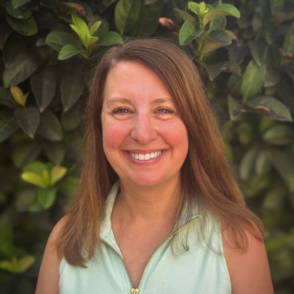 Smiling woman in a light green sleeveless top stands against lush green foliage.