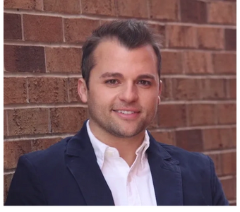 Smiling man in a navy blazer standing against a brick wall.