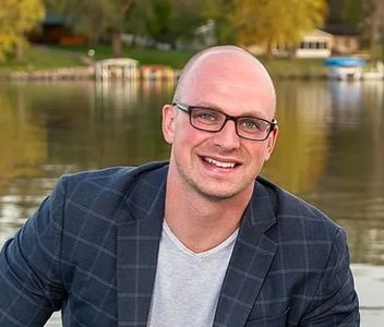 A smiling man with glasses and a blazer by a lakeside.