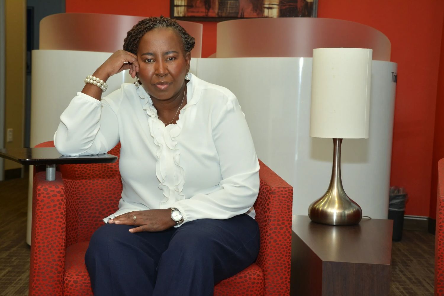 Woman in white blouse sitting on a red chair with a lamp beside her.