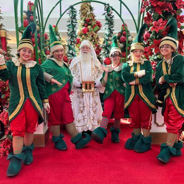 Santa Claus with five festive elves in a Christmas-themed setting.