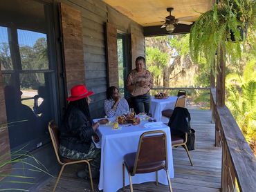 Three women enjoying a meal on a rustic wooden porch with nature around.