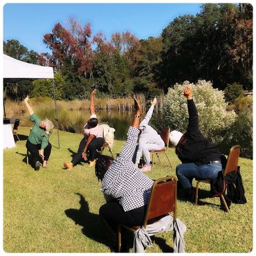 A group of people sitting on chairs outdoors doing a side stretch exercise.