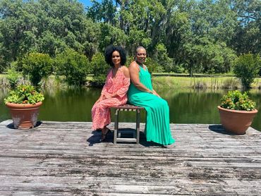 Two women in colorful dresses sit back-to-back on a bench by a serene pond.