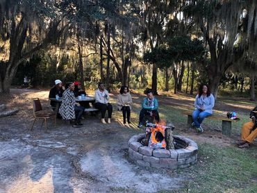 Group of people sitting around a campfire in a wooded area during daytime.