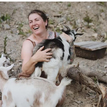 A joyful woman holding a small goat with two more goats nearby outdoors.